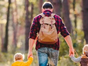 Papá agarrando a sus dos hijos en cada mano, caminando por un bosque soleado.