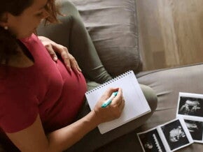 Primer mes de embarazo Mujer embarazada de sentada en un sofá, anotando nombres para bebés varones en un cuaderno con imágenes de ecografía a su lado.
