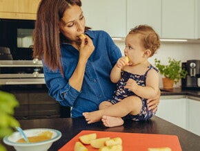 Identifica cuáles son las frutas ideales para iniciar esta nueva etapa de tu bebé. Mamá y bebé comiendo fruta en la cocina.