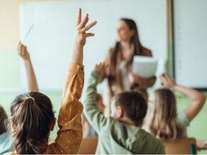 Salón de clase, profesora parada en frente al pizarrón y niños levantando la mano.