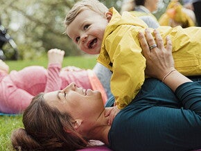 Mamá acostada sobre una colchoneta, al aire libre, cargando a su bebé y sonriendo, haciendo ejercicios para niños.