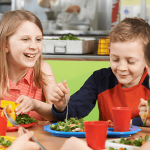 Niños comiendo en una cafetería escolar con platos de ensalada y pasta.