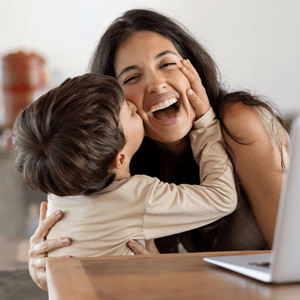 Niño dando un beso en el cachete a su madre sonriente. La mamá está sentada en frente de un ordenador portátil.