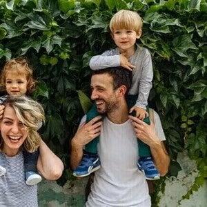 Dos adultos con dos niños sobre sus hombros, frente a una pared cubierta de hojas.