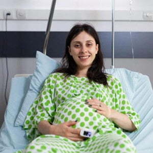 Mujer embarazada sonriendo, usando una bata de hospital y acostada en una camilla.