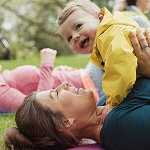Mamá acostada sobre una colchoneta, al aire libre, cargando a su bebé y sonriendo, haciendo ejercicios para niños.