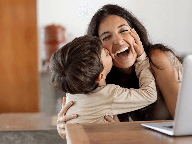 Niño dando un beso en el cachete a su madre sonriente. La mamá está sentada en frente de un ordenador portátil.