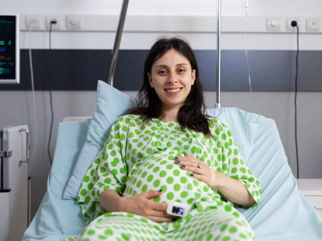 Mujer embarazada sonriendo, usando una bata de hospital y acostada en una camilla.