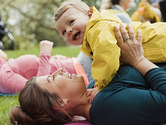 Mamá acostada sobre una colchoneta, al aire libre, cargando a su bebé y sonriendo, haciendo ejercicios para niños.