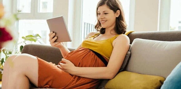 Mujer embarazada de sentada en un sofá, sonriendo y viendo en su tablet ideas de nombres para bebés varones. Mujer embarazada de sentada en un sofá, sonriendo y viendo en su tablet ideas de nombres para bebés varones.